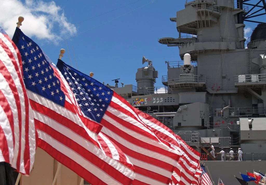 Image of a US Naval ship at port behind several billowing U.S. Flags; this picture is for an article discussing the risk to Navy NCOs and Officers of getting a page 13 for misconduct in the navy; show cause board lawyer